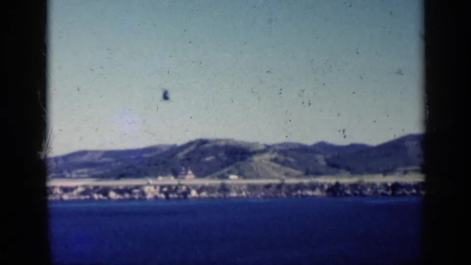1962 Cuba 8mm frame with a helicopter high over blue water, a distant coastal town, and green hills.