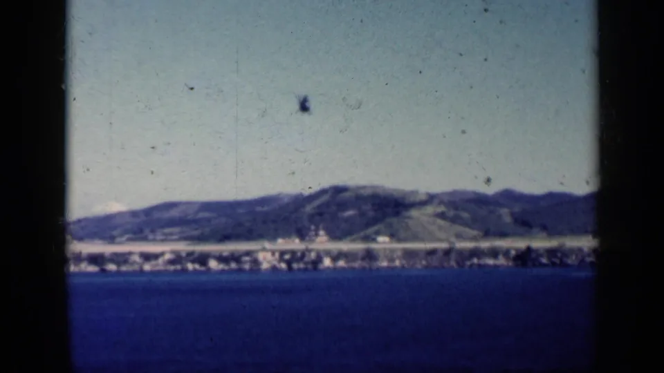1962 Cuba coastal home-movie frame showing open water, a distant shoreline, hills, and a helicopter shape above the horizon.