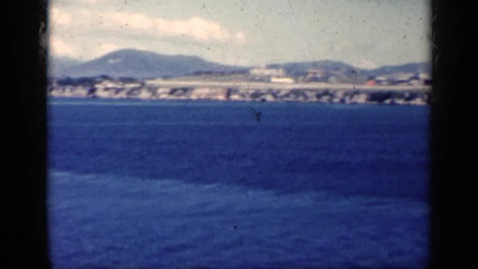 1962 Cuba 8mm scenic frame with wide blue water, distant hills, shoreline structures, and a light cloud above the horizon.