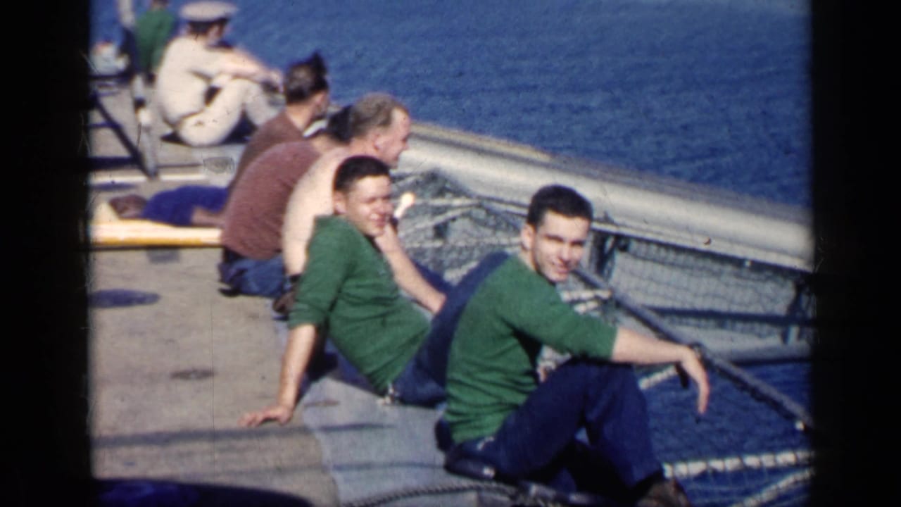 Several men sit on a ship deck beside railings with ocean behind them.