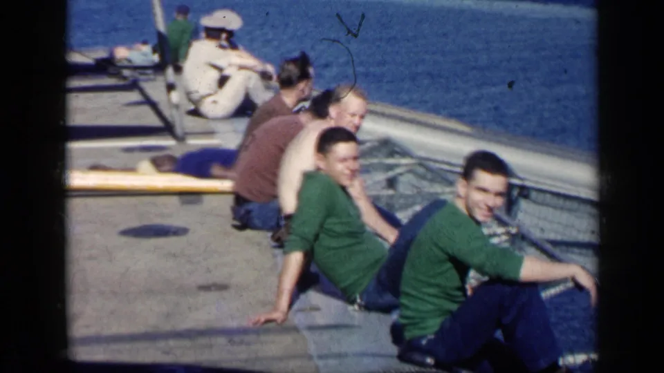 Several visible-faced men sit and look toward the camera on a ship deck, with ocean, railing, and a white-uniformed sailor behind them.