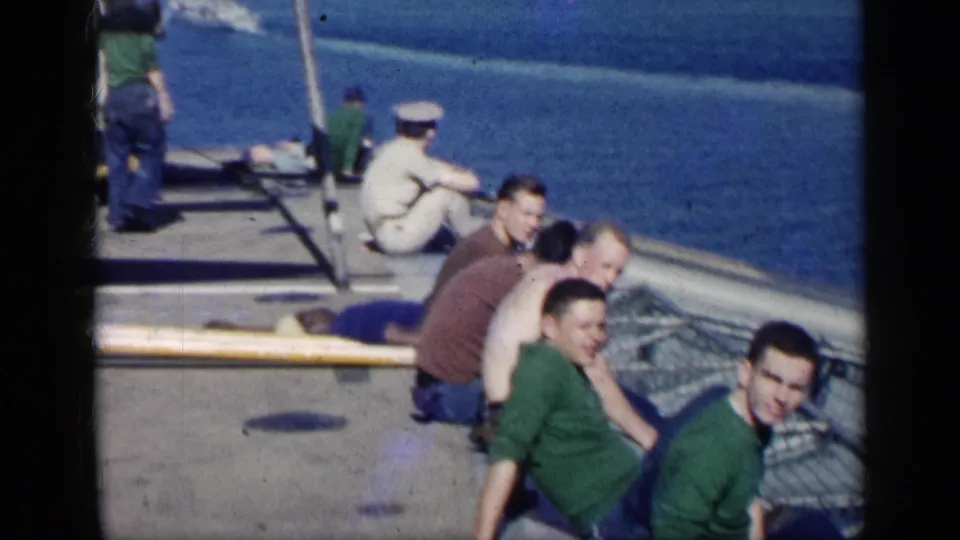 The home-movie frame shows men relaxing on a vessel deck near Cuba, with a white-uniformed sailor, blue water, and railings visible.