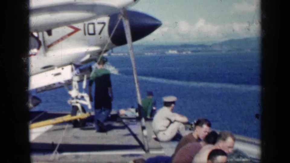 A shipboard scene shows part of an aircraft marked with numbers, sailors on deck, seated men, ocean, and a distant shoreline.