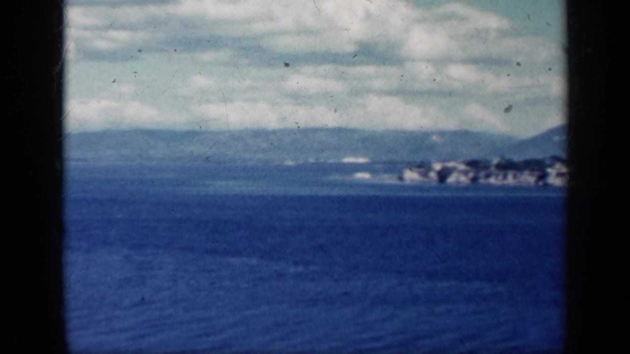 Blue water under cloudy sky with distant land and mountains on the horizon.