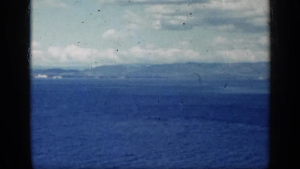 1962 Cuba coastal view across blue water toward a hazy mountain horizon beneath scattered clouds.