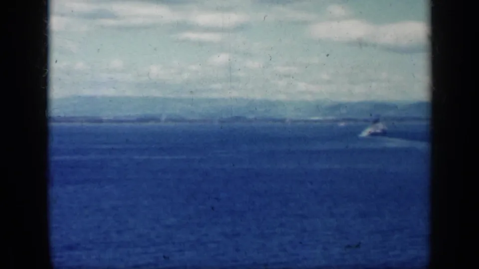 1962 8mm home-movie view over open water toward a distant shoreline, with cloudy sky and dark film gate edges.