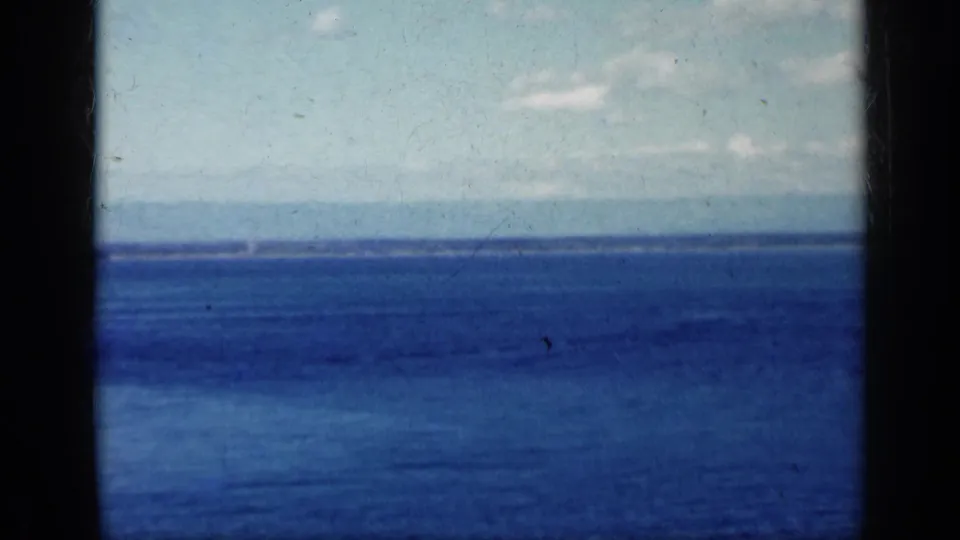 1962 8mm home-movie frame of blue ocean, cloudy sky, and a thin distant shoreline viewed from a moving vessel.