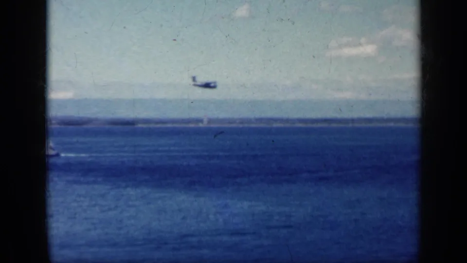 A small aircraft appears low over the water in 1962 8mm footage, with distant land and clouds behind it.