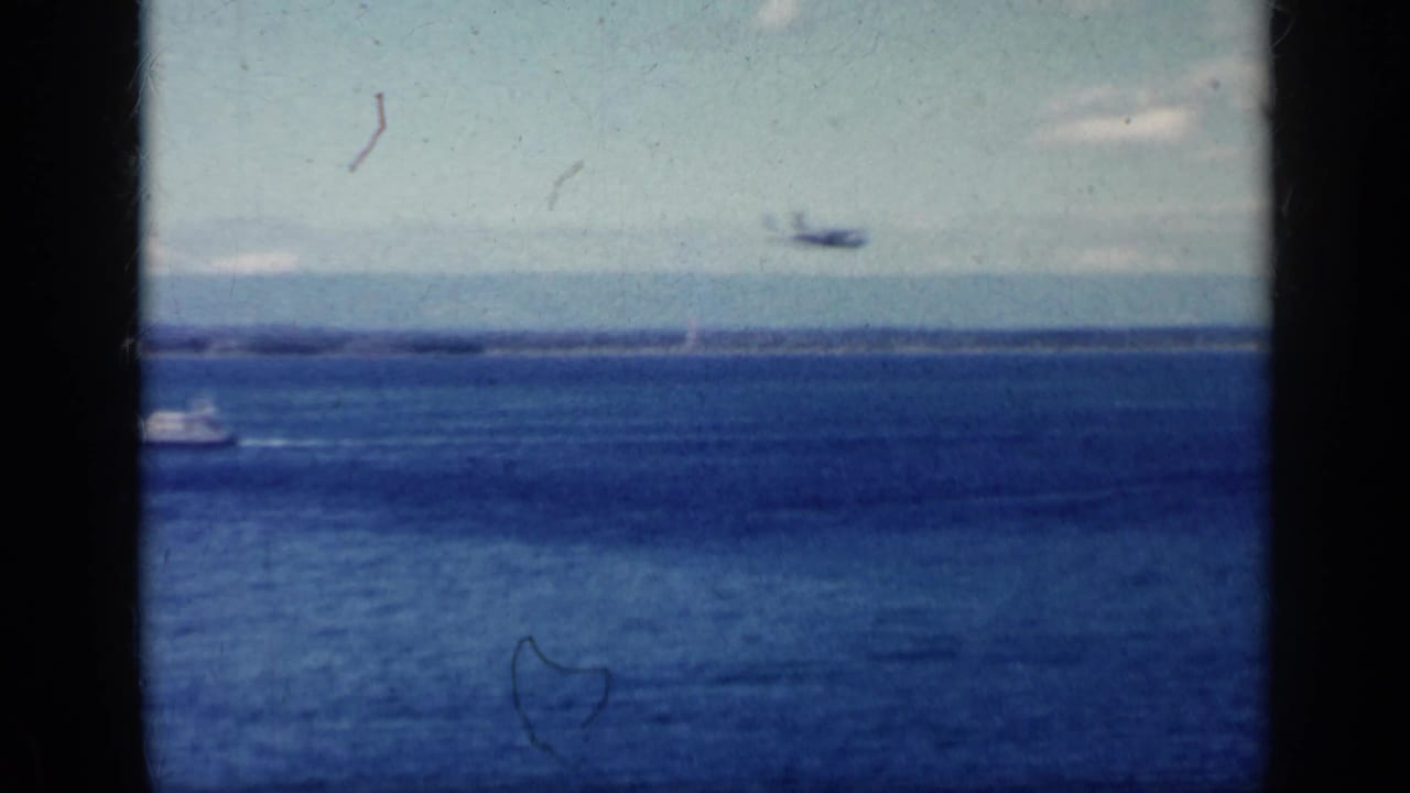 Blue ocean and distant shoreline seen through dark aircraft window framing, with a small plane in the sky.