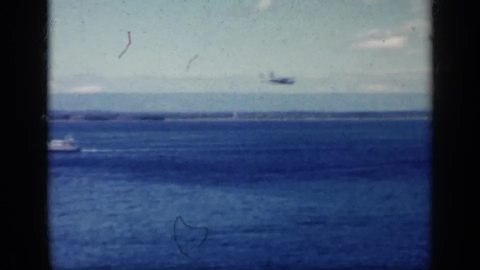 1962 Cuba home-movie frame showing ocean, pale sky, distant coast, and a small aircraft viewed through dark window divisions.