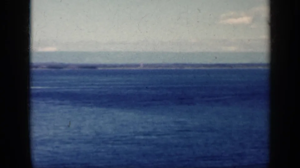 1962 Cuba-associated 8mm frame showing open blue water, pale clouds, and a tiny low-flying aircraft near the horizon.