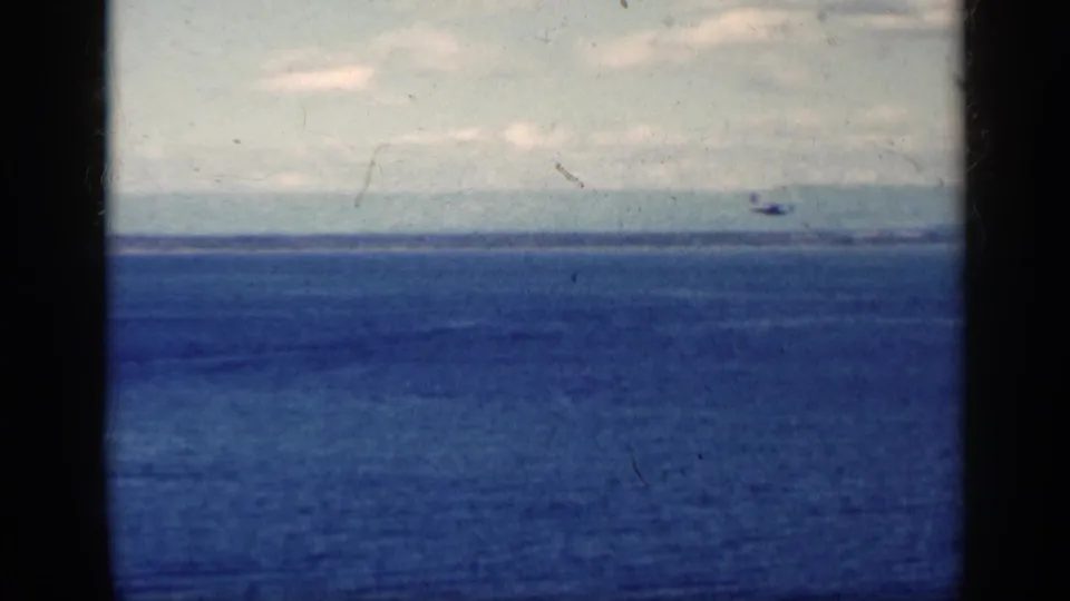 Cuba-associated 1962 8mm frame with a distant aircraft crossing above a blue sea under a pale, clouded sky.