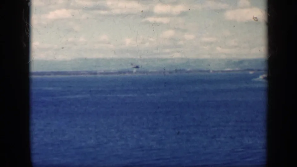 Cuba-associated 1962 8mm frame of a small low aircraft crossing a broad blue sea beneath light cloud cover.