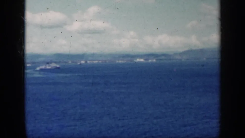 Vintage 8mm view of Cuba's coast, with blue water in the foreground, a ship at left, hazy hills, and puffy clouds.