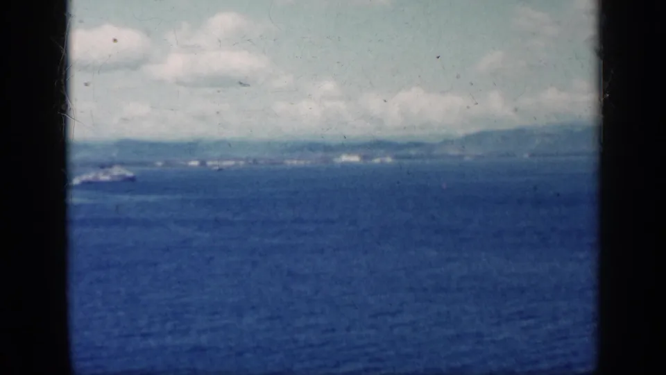1962 Cuba shoreline scene with a small ship near the left edge, deep blue water, distant land, and white clouds in soft 8mm color.