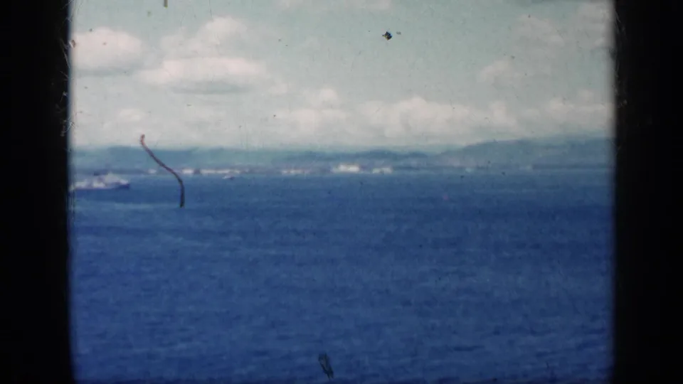 1962 Cuba-linked 8mm frame showing open blue water, a low distant shoreline, small ships, cloudy sky, and dark film gate edges.