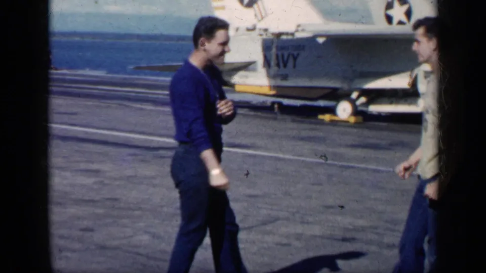 1962 Cuba-linked 8mm frame of two adult men conversing on a military tarmac beside a Navy-marked jet aircraft.
