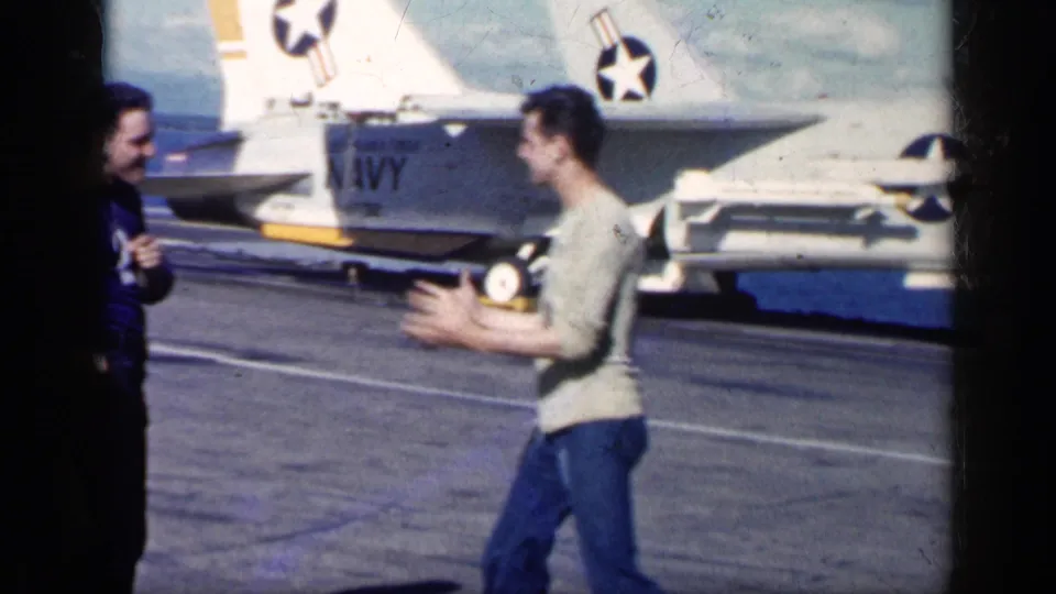 Cuba-linked 1962 8mm frame showing two men on a military airfield beside a Navy-marked jet, with sea visible beyond the tarmac.