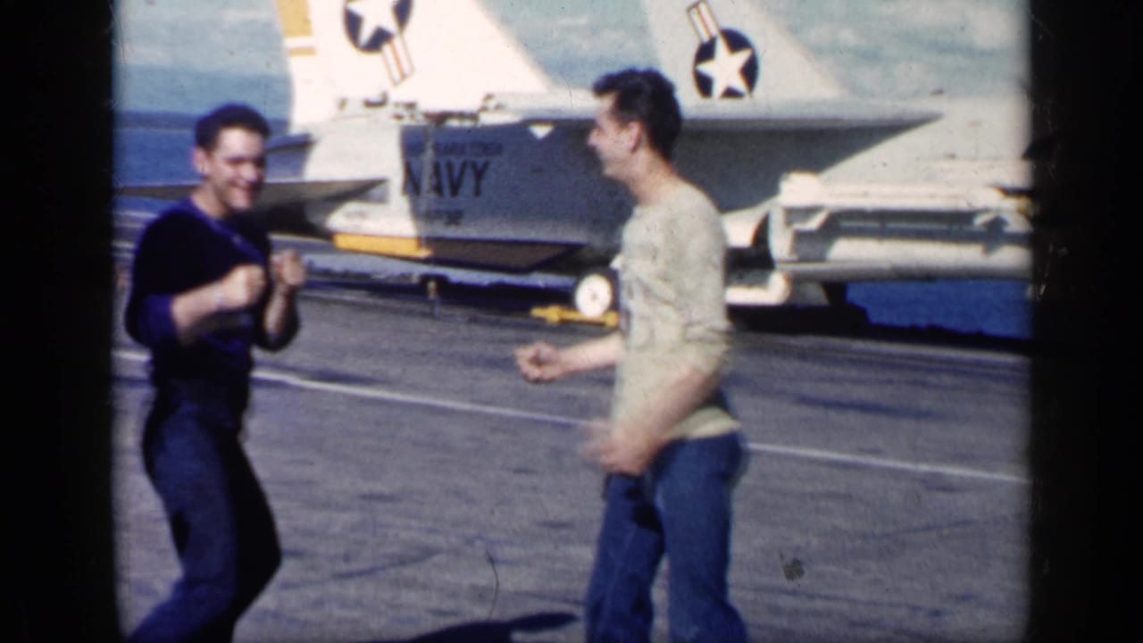 Two men playfully square off on an aircraft carrier deck beside a Navy jet tail.