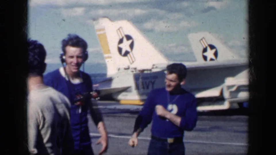 Three young men gather on the carrier deck near Navy aircraft markings, one wearing headphones and a blue uniform-style shirt.