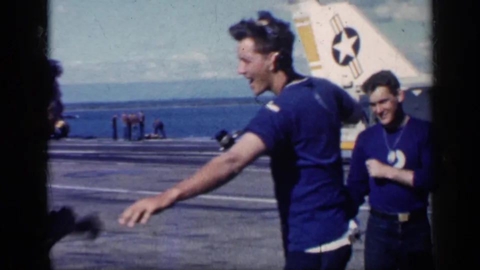 A man in blue walks or gestures across the flight deck, with another smiling man and Navy aircraft tail markings in the background.