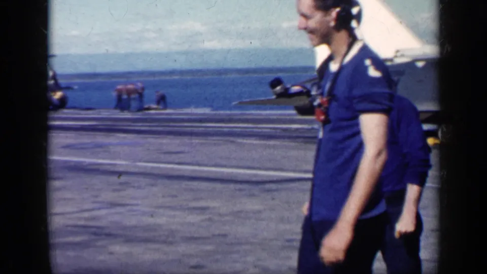 A smiling Navy man wearing headphones stands on the carrier deck, with ocean, deck lines, and another man partly visible behind him.
