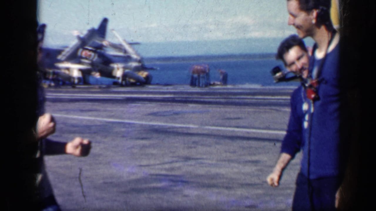 Men on an aircraft carrier deck with parked naval aircraft and ocean behind them.
