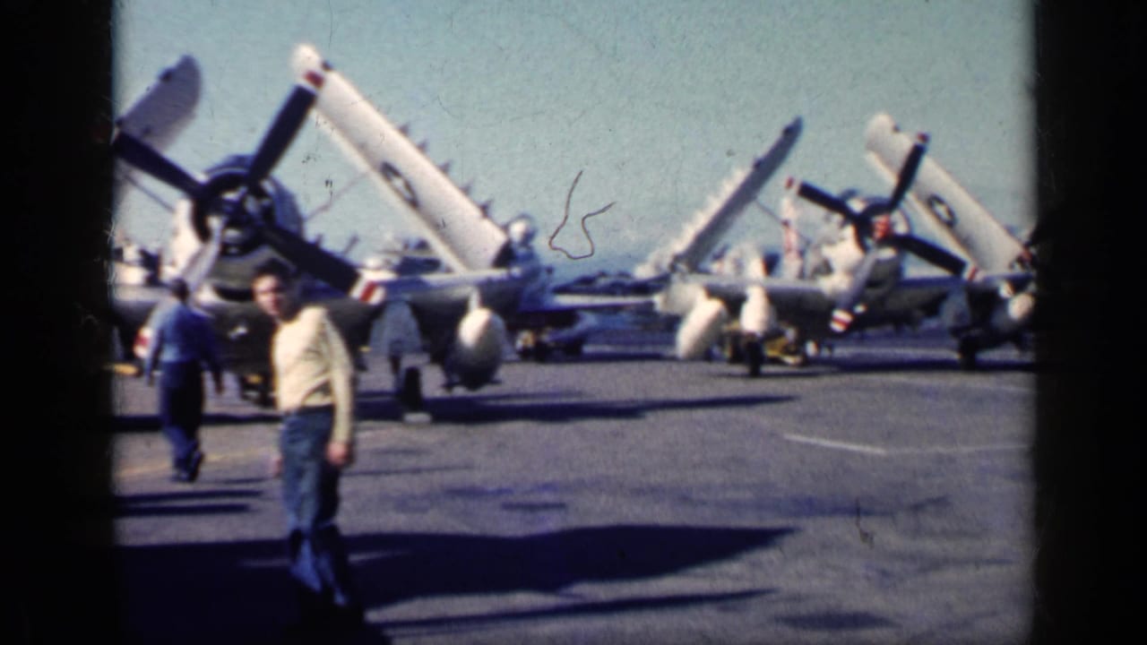 Man standing on an aircraft carrier deck with several propeller planes behind him.