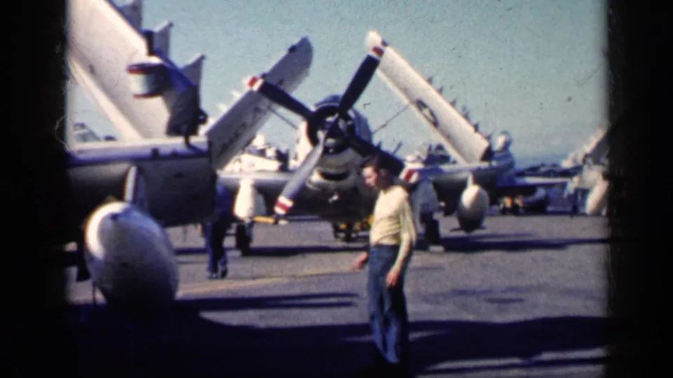 A man moves across a carrier deck in the supplied Cuba 1962 clip, with folded-wing propeller planes and other people nearby.