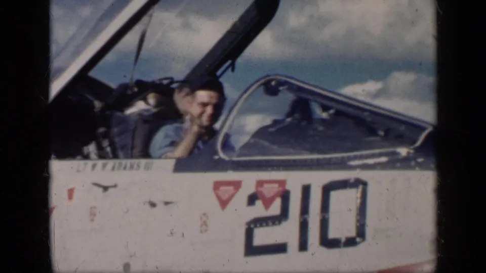 Smiling pilot seated in an open military jet cockpit on an aircraft carrier deck, with number 210 visible, in the 1962 Cuba clip context.
