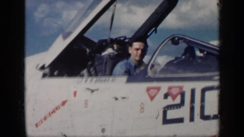 1962 Cuba-associated 8mm frame of a military pilot in a blue flight suit seated in an open U.S.-marked jet cockpit.
