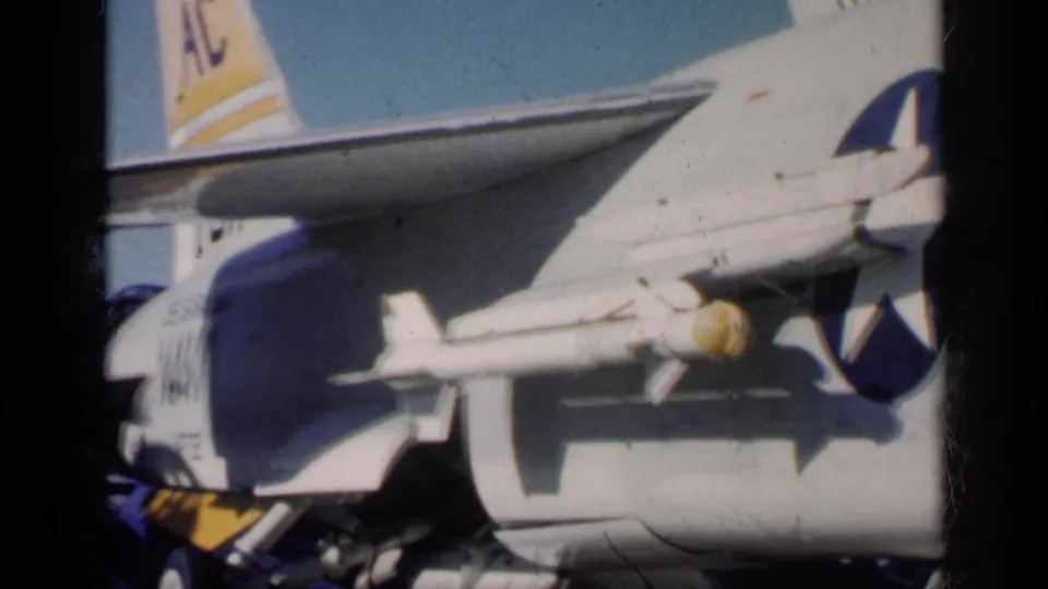 Final 8mm frame shows the U.S.-marked aircraft with underwing ordnance and partial tail markings, with another aircraft section or tail visible behind it.