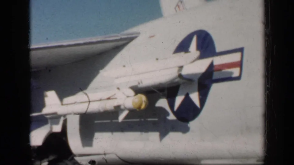 Close 8mm view of the Navy aircraft side, showing the U.S. roundel, wing structure, and a yellow-nosed rocket under the wing.