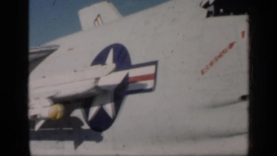 1960s Navy aircraft close-up showing the U.S. roundel, underwing rocket, and red stencil text on the fuselage in faded 8mm color.