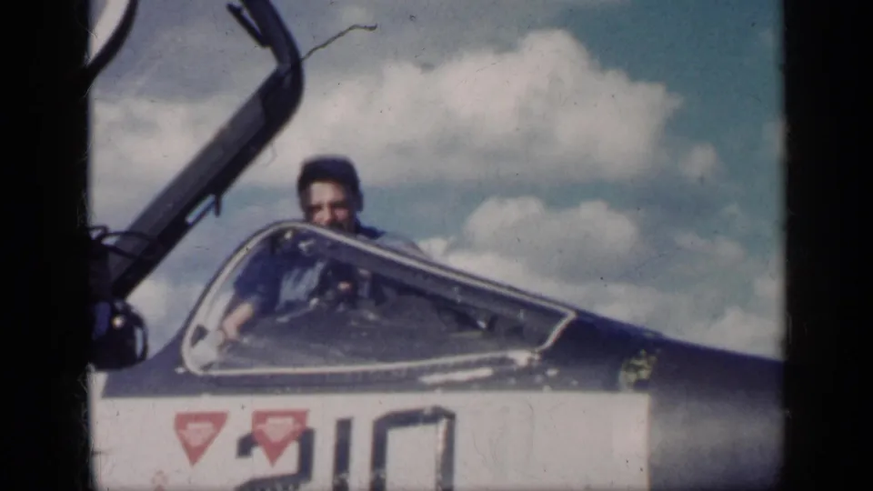 The man remains partly visible behind the aircraft cockpit windscreen, with painted markings on the fuselage and clouds behind him.