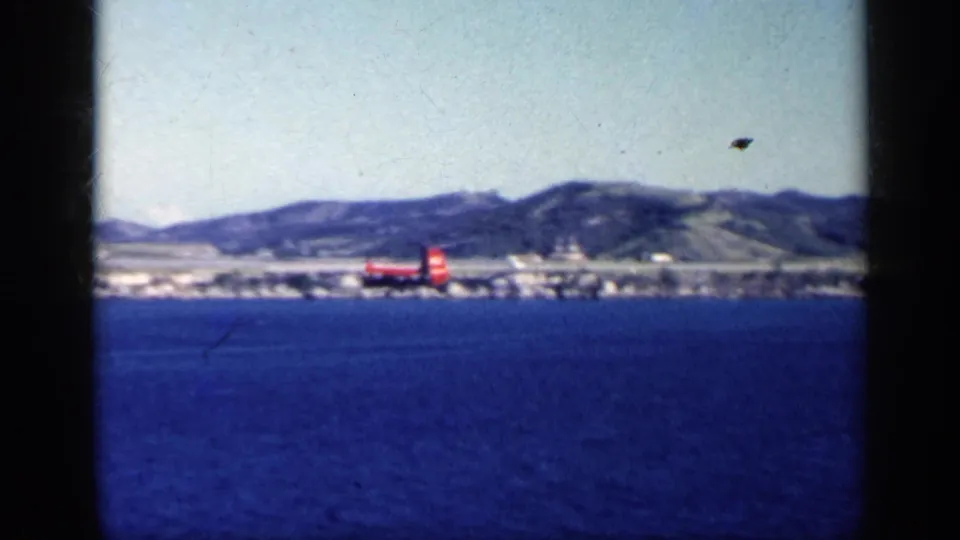 A red helicopter flies low over blue water near a distant Cuban shoreline and low hills in 1962 8mm footage.