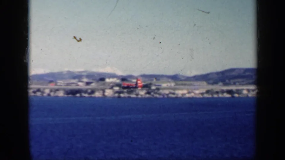 The red helicopter is seen over water near a distant Cuban coastal settlement and hills, with soft 8mm color and dust marks.
