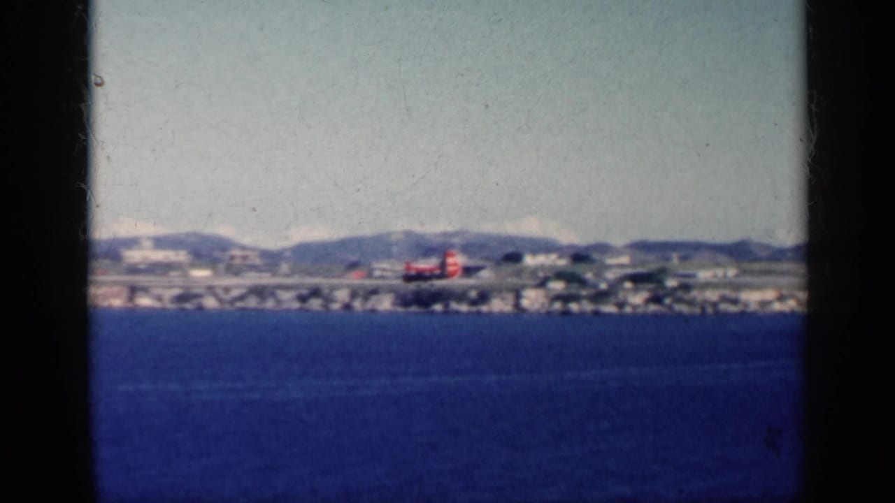 1962 Cuba home-movie frame showing a small red aircraft on a distant coastal runway beyond blue ocean water and low mountains.