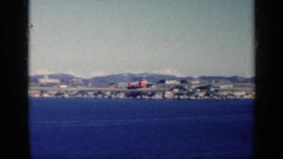 1962 Cuba home-movie frame showing a small red aircraft on a distant coastal runway beyond blue ocean water and low mountains.