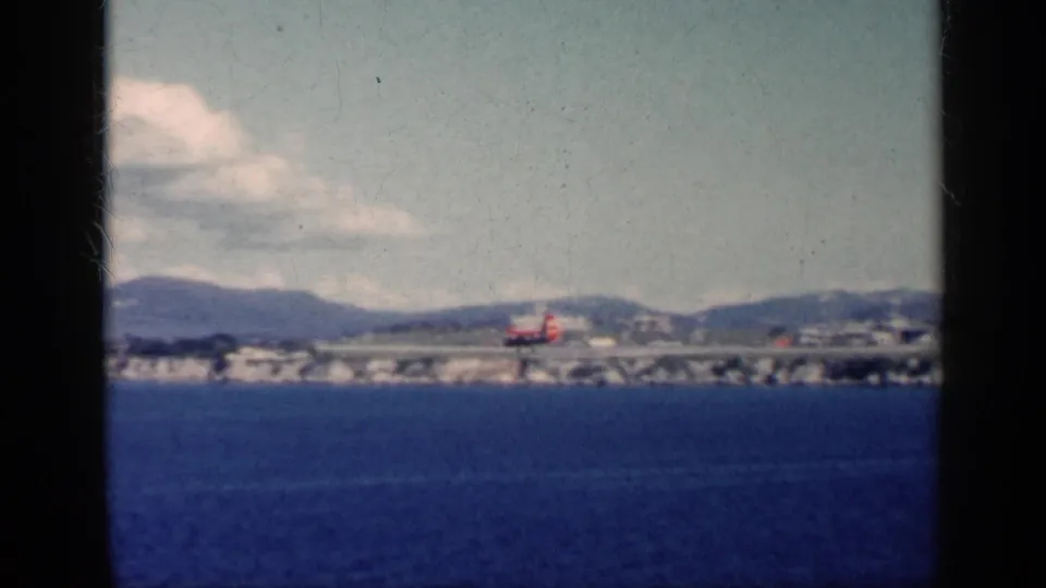 A distant 1962 Cuba coastal airfield view, with a red aircraft near the runway, ocean in the foreground, and hazy hills beyond.