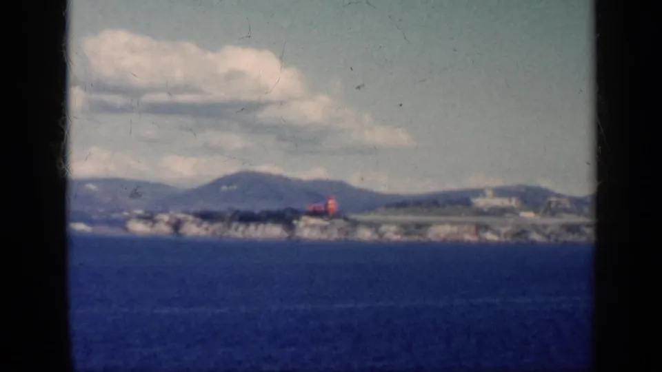 Soft 8mm frame of a small red aircraft on a distant Cuban coastal runway, with ocean foreground, low buildings, hills, and clouds.