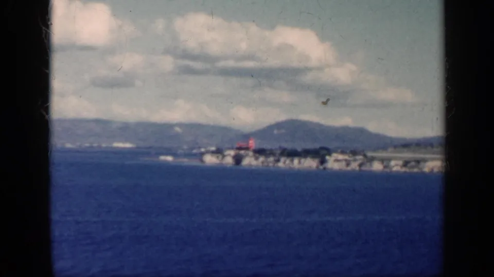 Final 1962 Cuba frame showing the red aircraft at a far coastal runway, blue water below, and mountains under a cloudy sky.