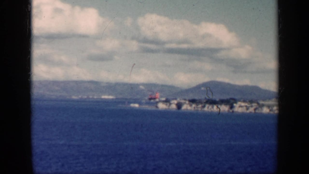 1962 Cuba 8mm frame showing blue water, distant hills, cloud cover, and a small aircraft near the shoreline.