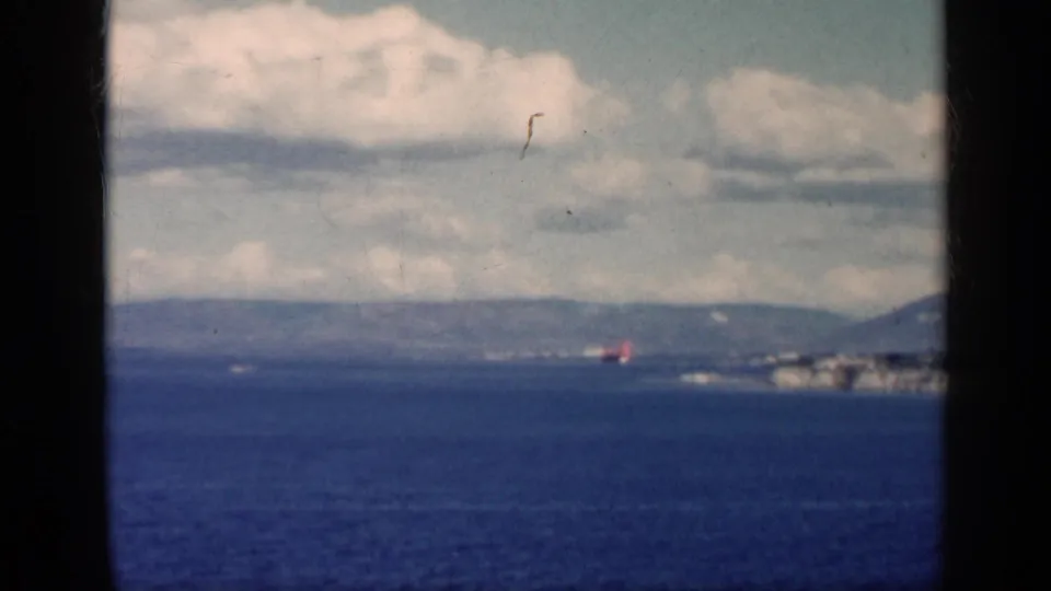 1962 Cuba 8mm frame with open water, distant coastline, low hills, cloud bands, and a small red shoreline detail.