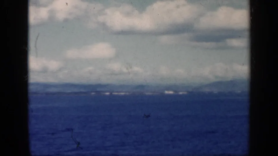 1962 Cuba 8mm frame of open water, distant shore, low hills, cloudy sky, and dark gate borders from the film scan.