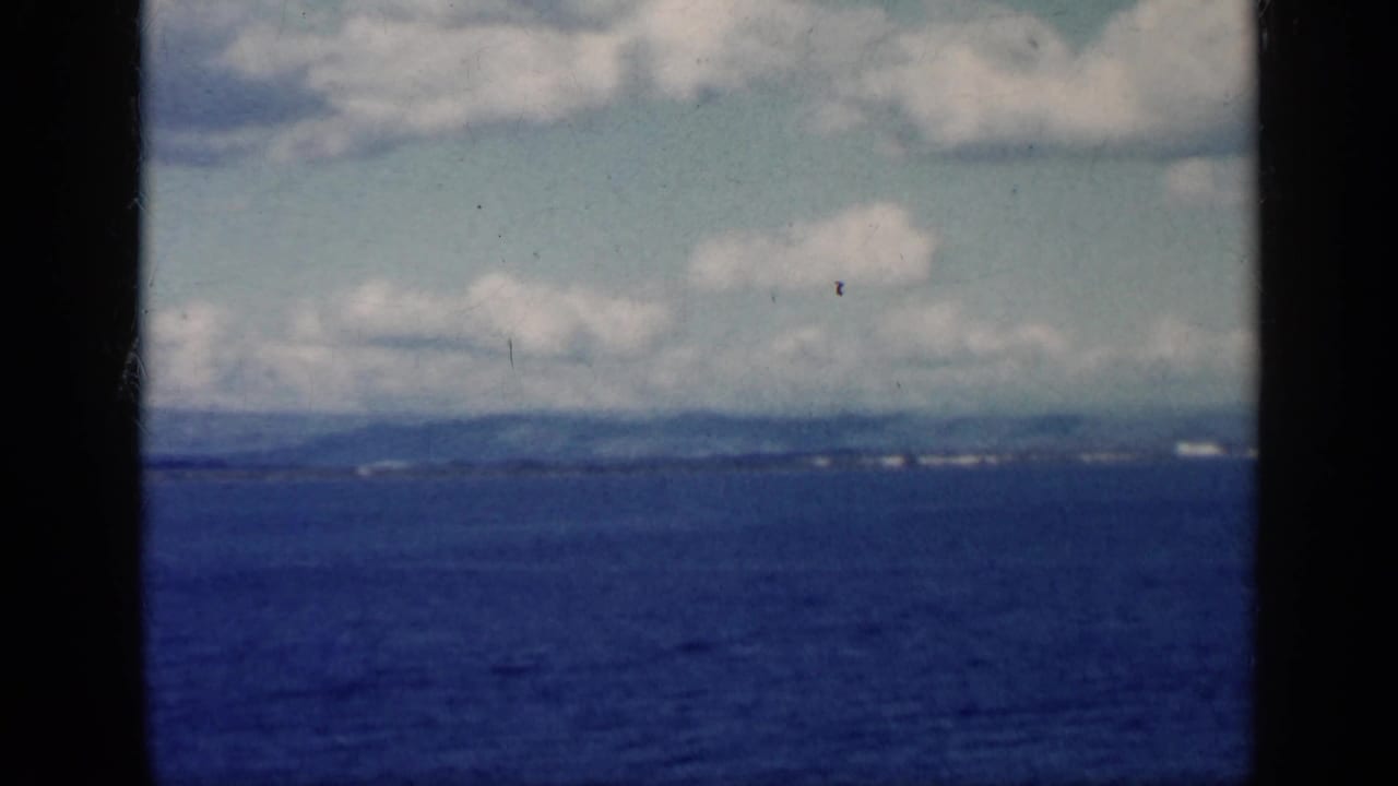 1962 Cuba-labeled 8mm frame showing blue sea, distant shoreline, clouds, and dark aircraft-carrier structure at the edges.
