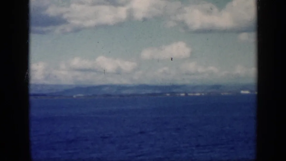 1962 Cuba-labeled 8mm frame showing blue sea, distant shoreline, clouds, and dark aircraft-carrier structure at the edges.