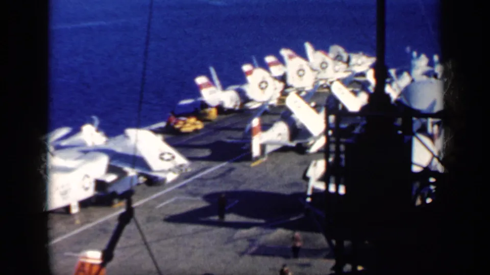 A 1962 Cuba-labeled carrier-deck frame shows parked Navy jets, deck shadows, rigging, blue sea, and small crew figures near the lower edge.