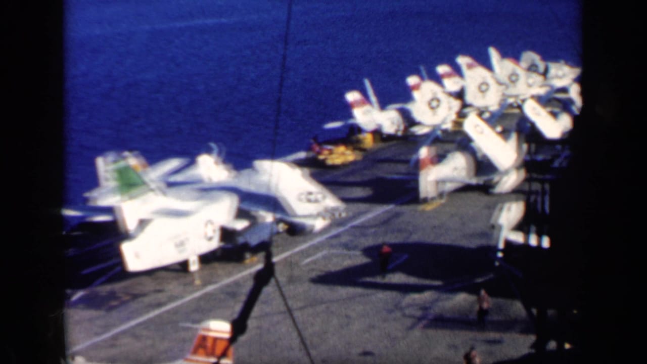 1962 Cuba-era 8mm frame showing white U.S. Navy aircraft lined on a carrier deck with ocean beyond and deck shadows.