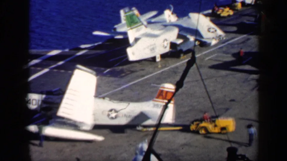 High-angle 8mm view of U.S. Navy aircraft on a carrier deck near Cuba in 1962, with crew figures and a yellow deck vehicle visible.